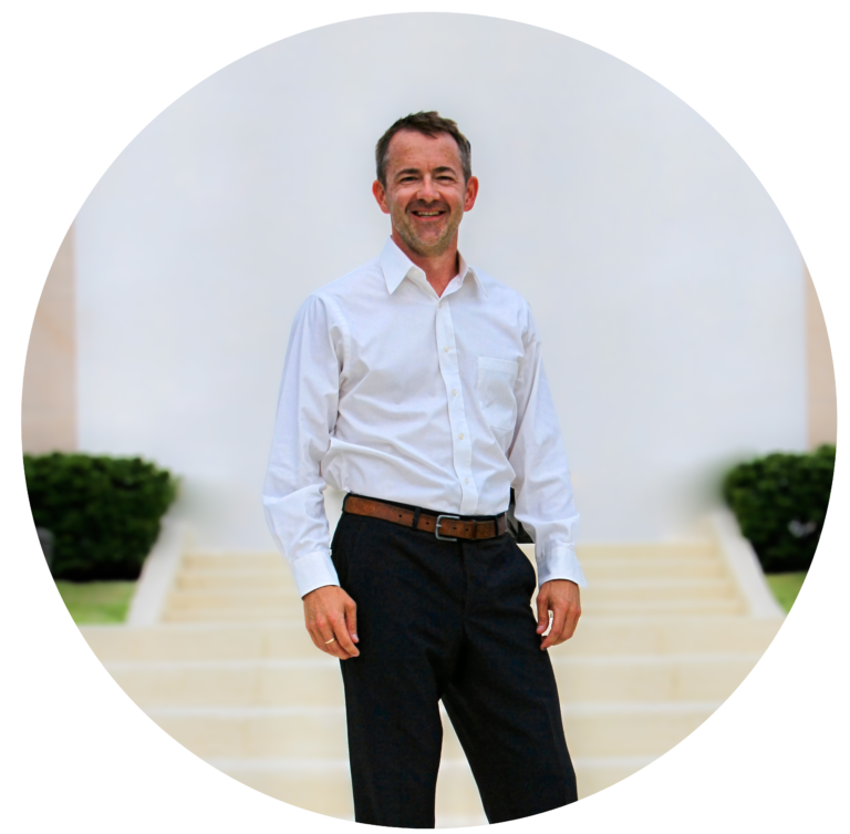 Confident Man In A White Shirt And Dark Trousers, Smiling Outdoors Against A Serene Backdrop.